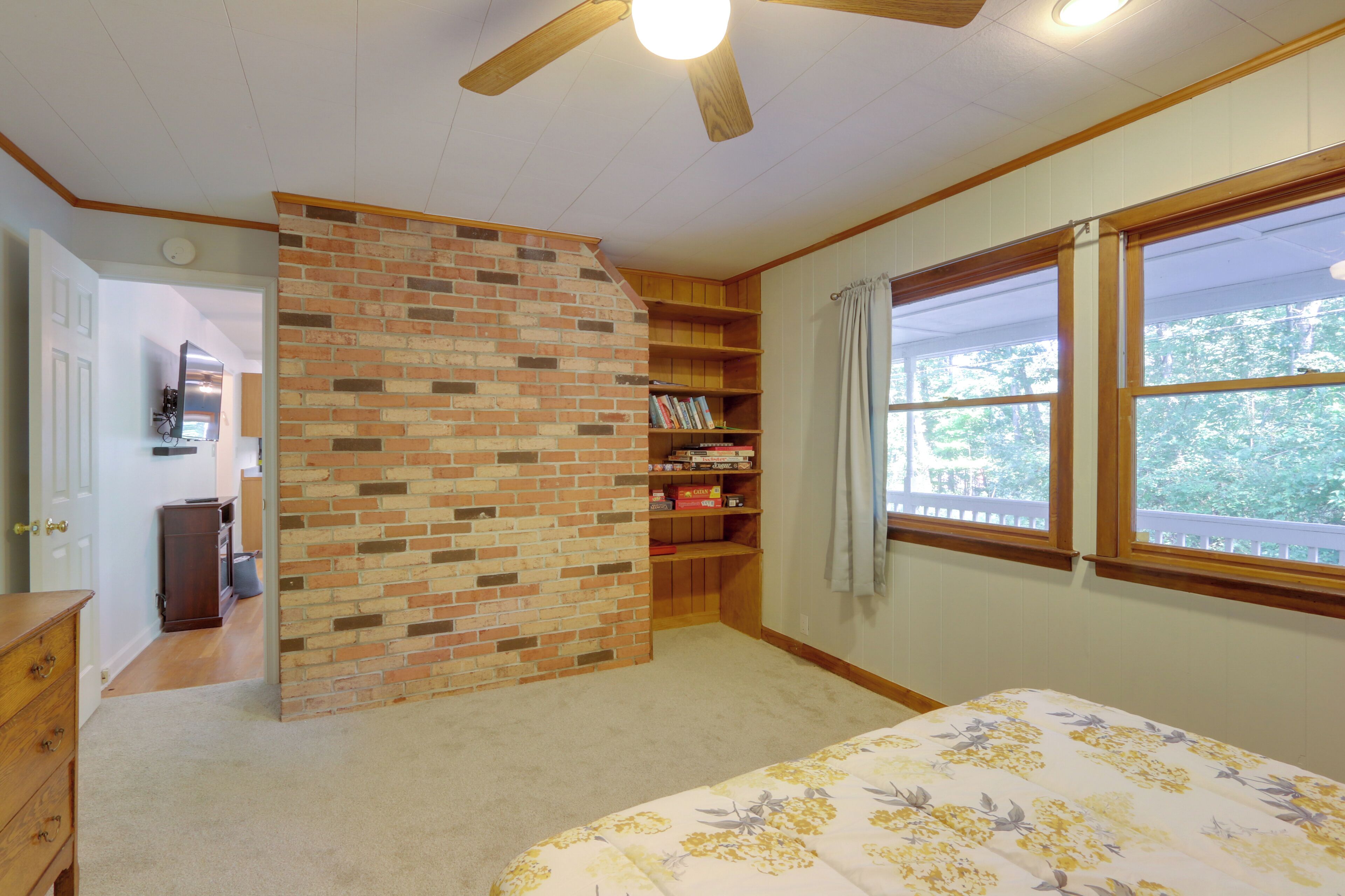 A bedroom with a brick accent wall, a wooden bookshelf, a bed with floral bedding, and a window with a view of trees.