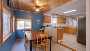 A spacious dining area with a wooden table and chairs, a kitchen with wooden cabinets, and a large window providing natural light. - Harpers Ferry Cabin - Walk to Private Lake! (Harpers Ferry)