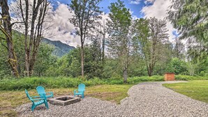 A gravel path leading to a seating area with two chairs and a fire pit, surrounded by dense forest and a mountain in the background.