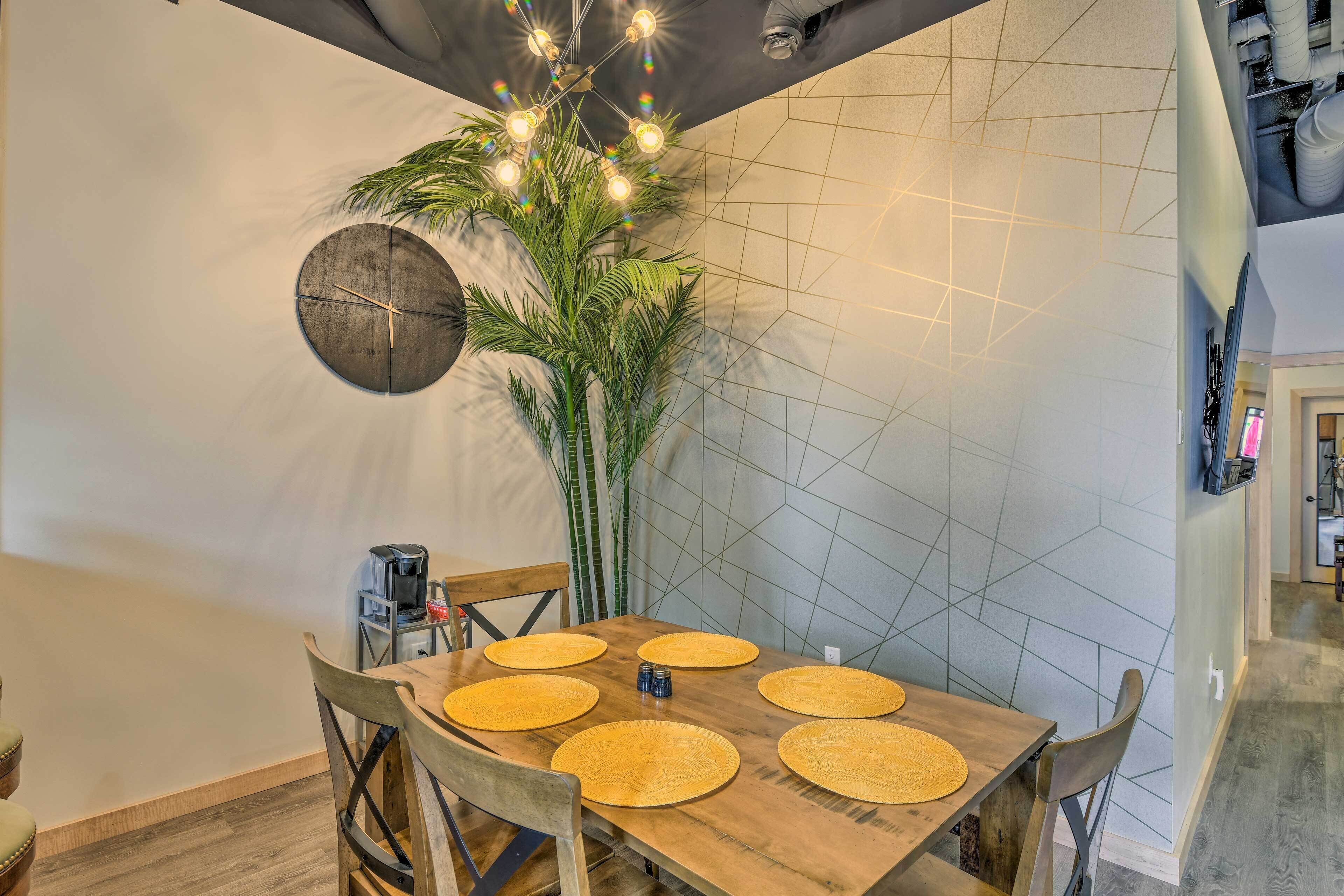 Un área de comedor con una mesa de madera, seis individuales y sillas. Hay una cafetera y una planta sobre la mesa.