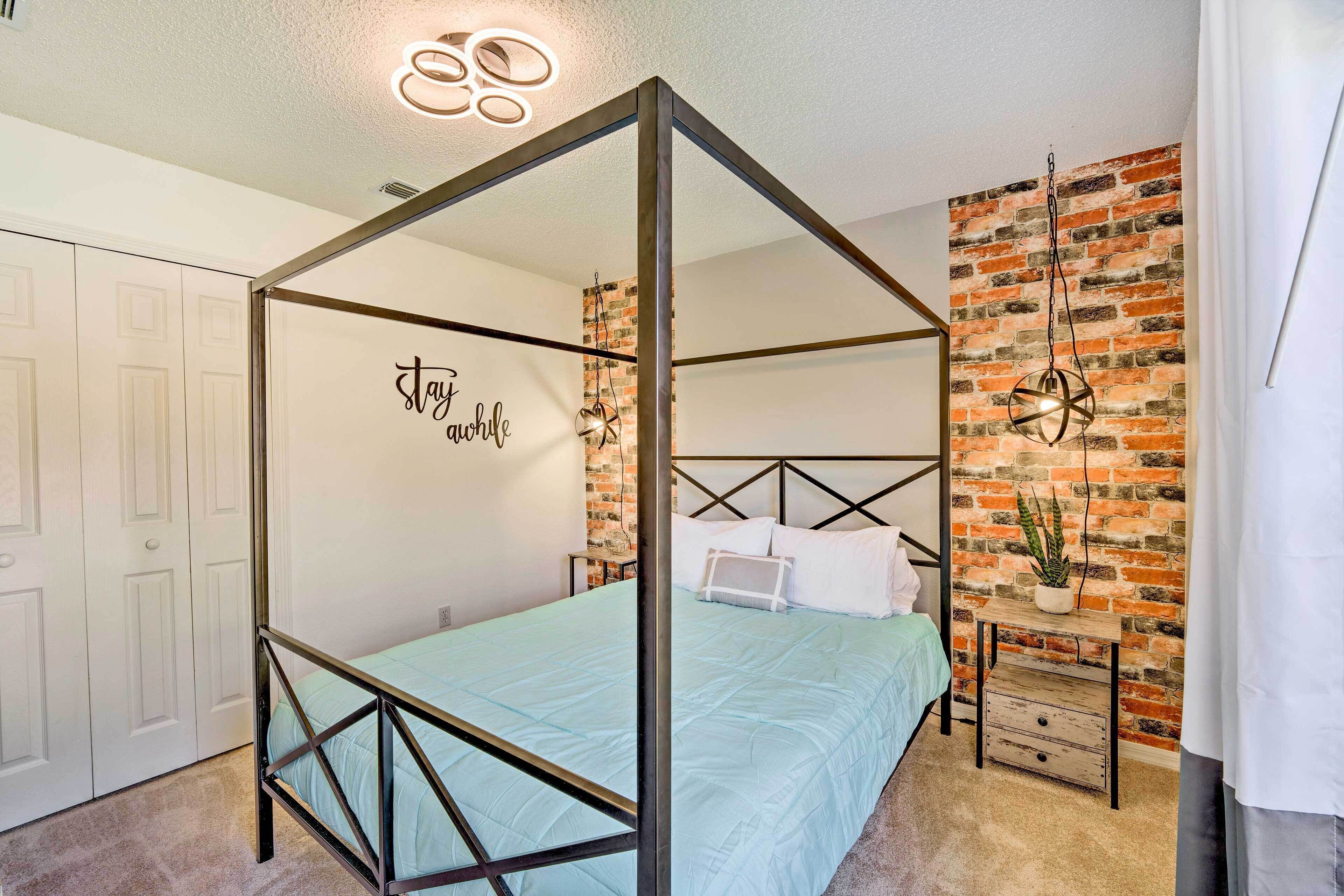 A four-poster bed with a light blue bedspread, a white wall with a 'stay awhile' sign, a brick wall, and a small wooden nightstand with a plant.