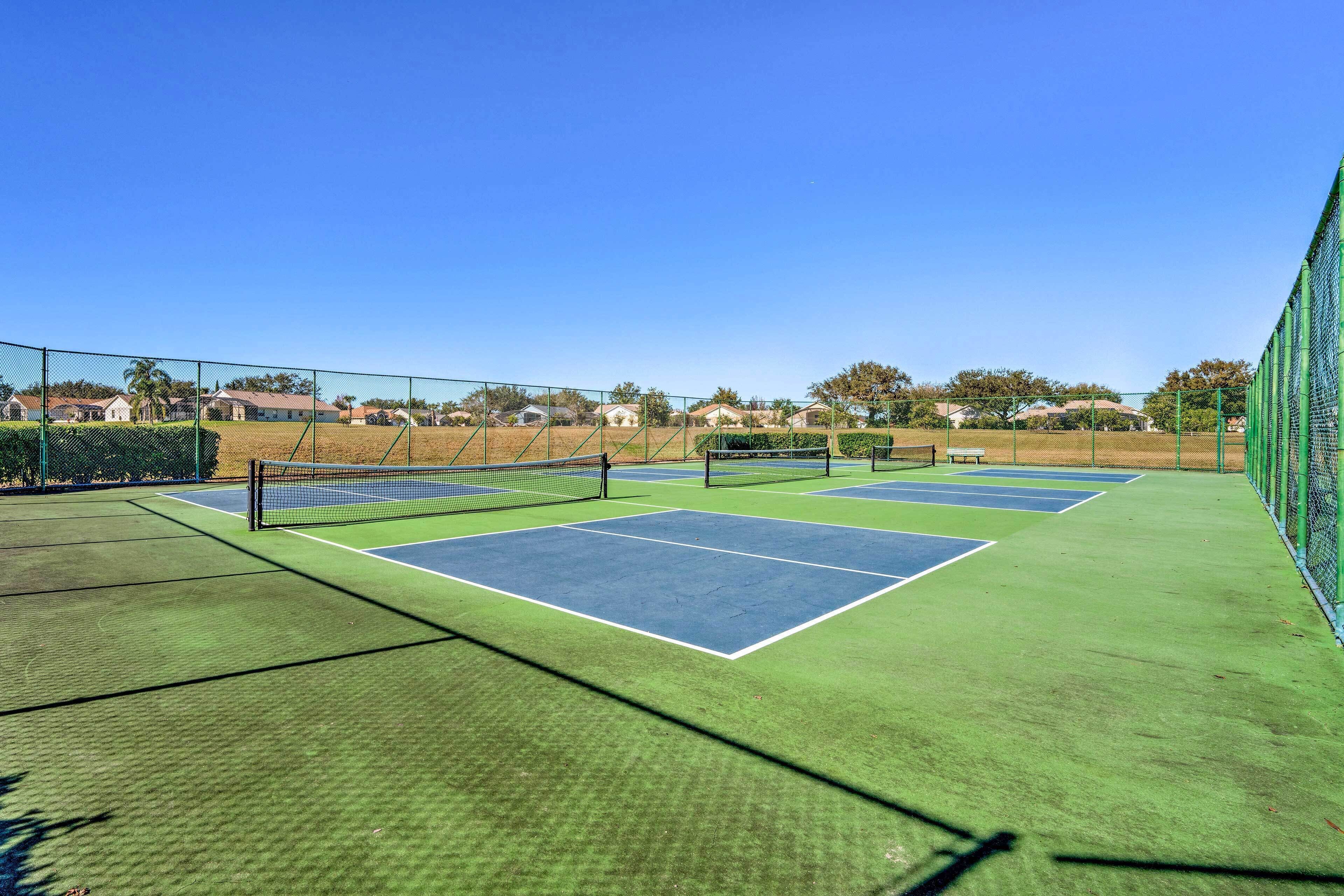 Canchas de tenis múltiples, rodeadas por una valla, y casas al fondo.