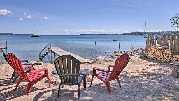 Uma área de estar à beira-mar, com cadeiras Adirondack e uma pequena mesa, com vista para águas calmas, barcos e uma linha de costa distante.