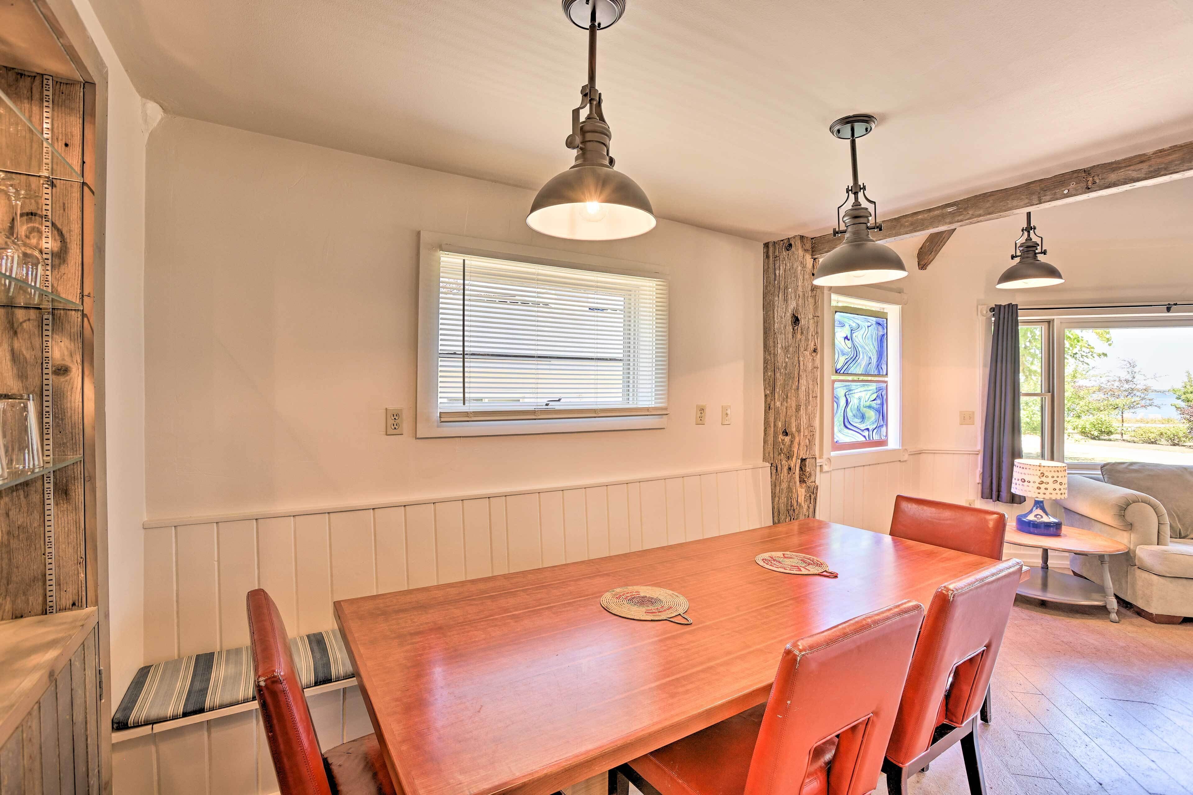 A dining area with a wooden table, chairs, pendant lights, a bench, and a window with blinds.