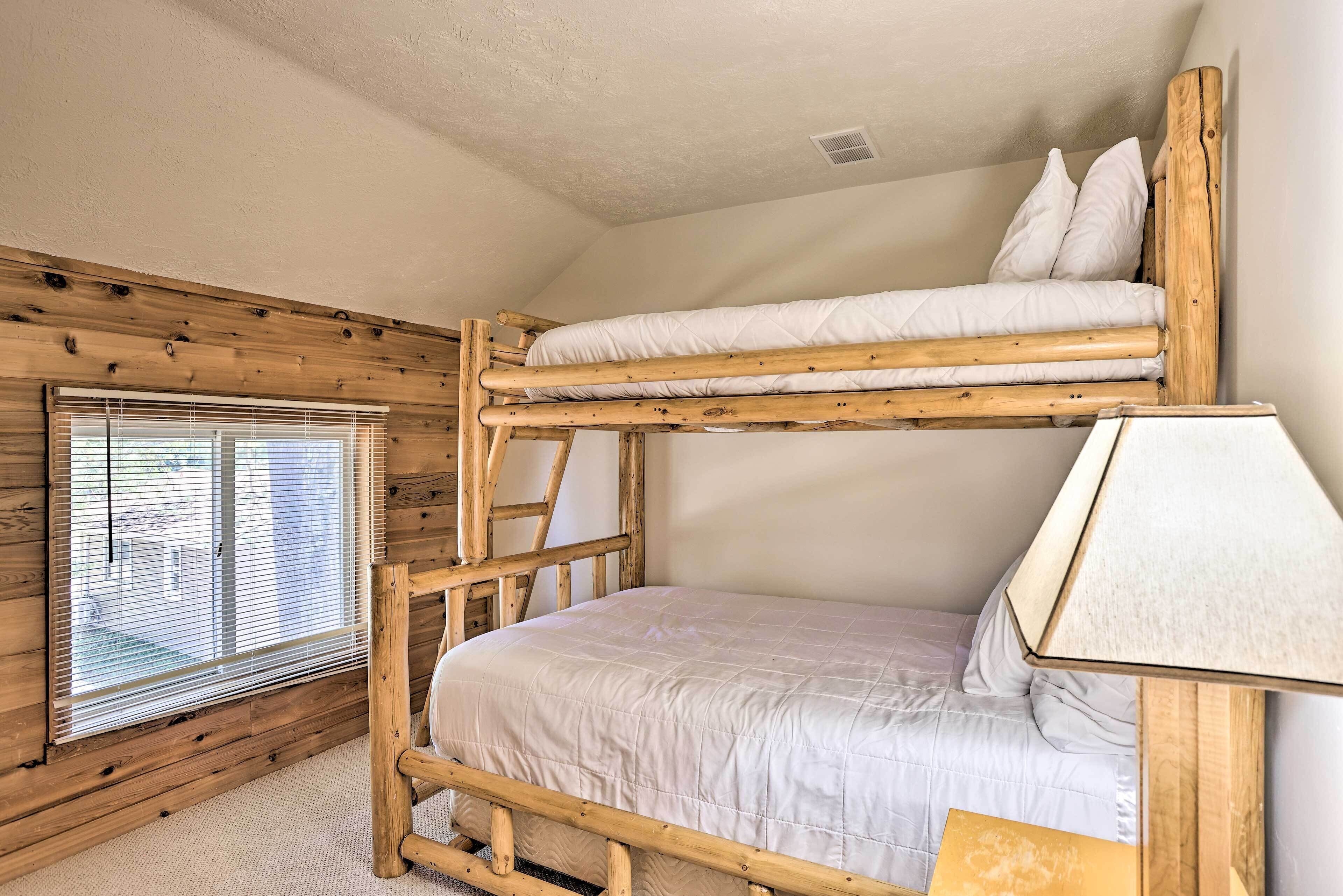 A bunk bed with a wooden frame and white bedding, a window with blinds, and a lamp on the side.