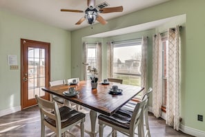 Un coin repas avec une table en bois, des chaises blanches, un ventilateur de plafond et de grandes fenêtres avec des rideaux.