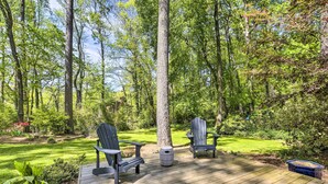 Une terrasse en bois avec deux fauteuils Adirondack et une petite table ronde, le tout entouré d’une végétation luxuriante et d’arbres.