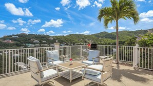 Un balcón con mobiliario exterior blanco, una palmera y vistas a un paisaje montañoso con casas.