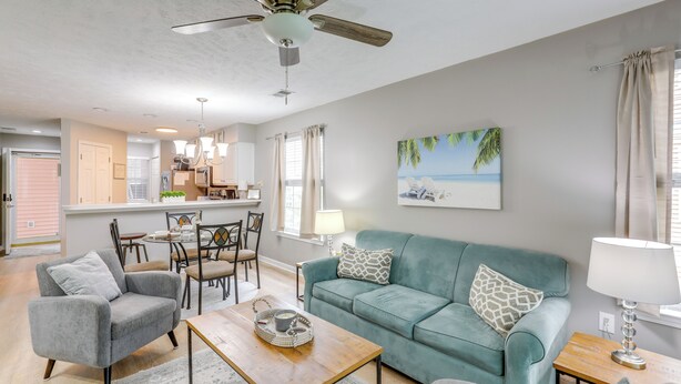 A spacious living room with a teal sofa, a grey armchair, and a wooden coffee table. There is a ceiling fan, a chandelier, and a painting of a beach scene on the wall. - 'southern Charm' Condo w/ Pool ~ 4 Mi to Broadway! (Myrtle Beach)