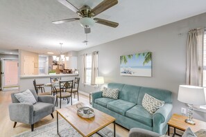 Uma sala de estar ampla com um sofá azul-turquesa, uma poltrona cinza e uma mesa de centro de madeira. Há um ventilador de teto, um lustre e um quadro com uma paisagem de praia na parede.