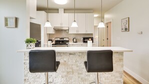A modern kitchen with white cabinets, a central island with two black bar stools, pendant lighting, and a stone backsplash.