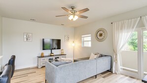 A modern living room with a grey sofa, a flat-screen TV, a ceiling fan, and a large window with curtains.