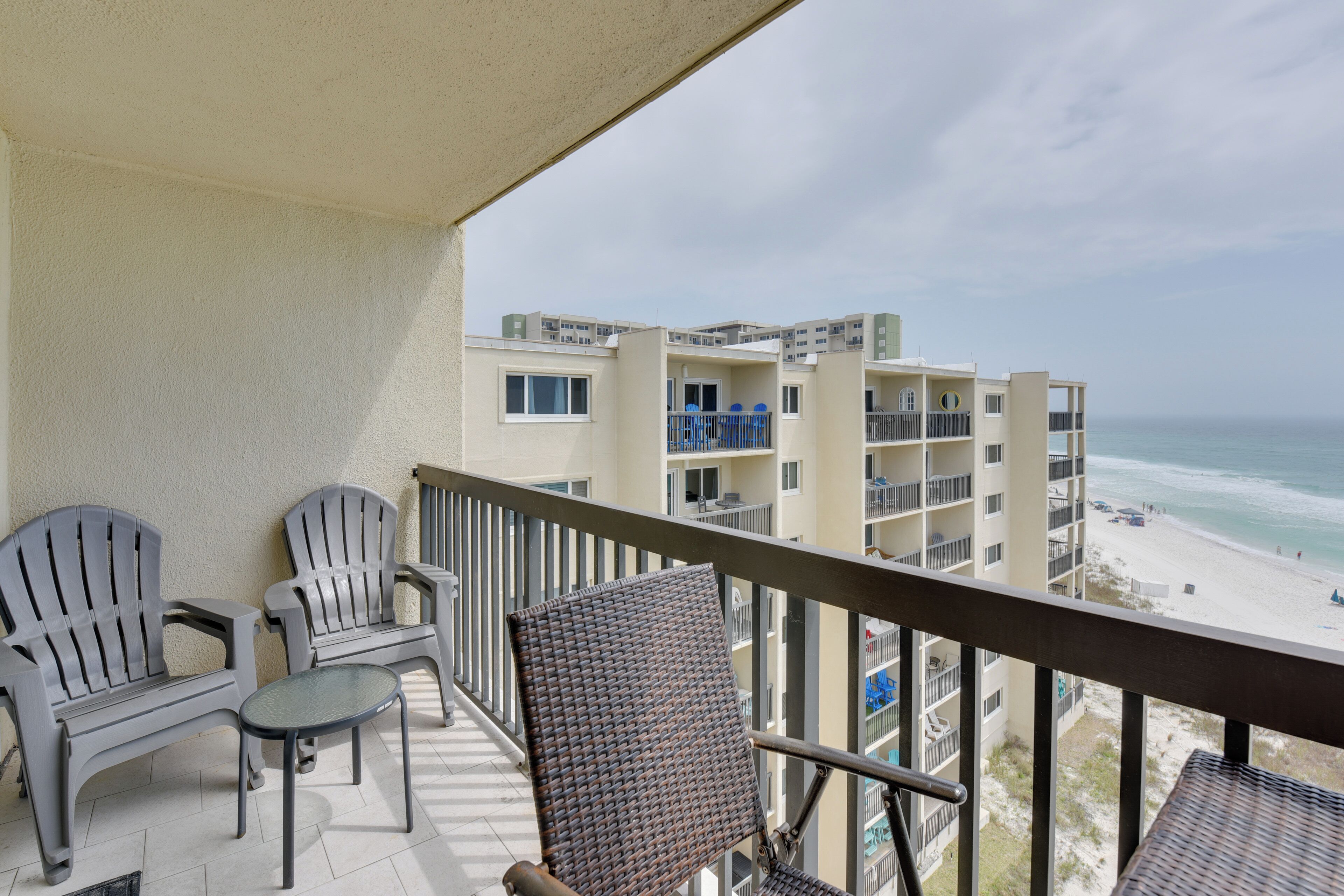 Un balcón con dos sillas Adirondack, una mesita y una silla de mimbre. El balcón ofrece vistas a una playa con un edificio de varios pisos y el océano.