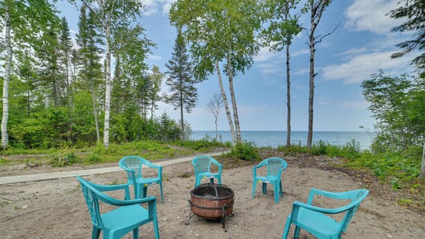 Een strandsetting met een vuurkuil en vier turquoise stoelen in een kring, omringd door bomen en met uitzicht op zee.
