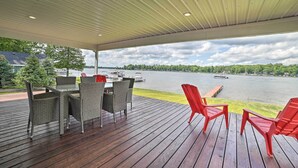 Une terrasse extérieure couverte, aménagée avec des meubles en osier et des chaises rouges, qui offre une vue sur un lac où l’on peut apercevoir des bateaux et un quai.
