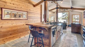 A spacious kitchen with wooden cabinetry, a central island with stools, and a large window providing natural light. - Hot Tub & Mtn Views: Waterfront Grand Lake Cabin! (Grand Lake)