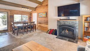 A cozy living room with a fireplace, wooden walls, a dining area, and a bookshelf. - Hot Tub & Mtn Views: Waterfront Grand Lake Cabin! (Grand Lake)