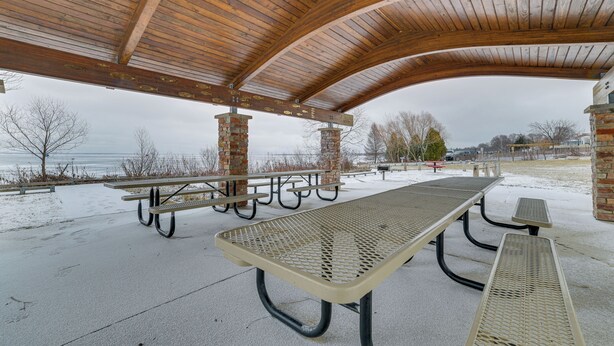 A covered picnic area with picnic tables and benches, a view of the water, and a brick building in the background. - Historic Houghton Lake Getaway Near Shoreline (Houghton Lake)