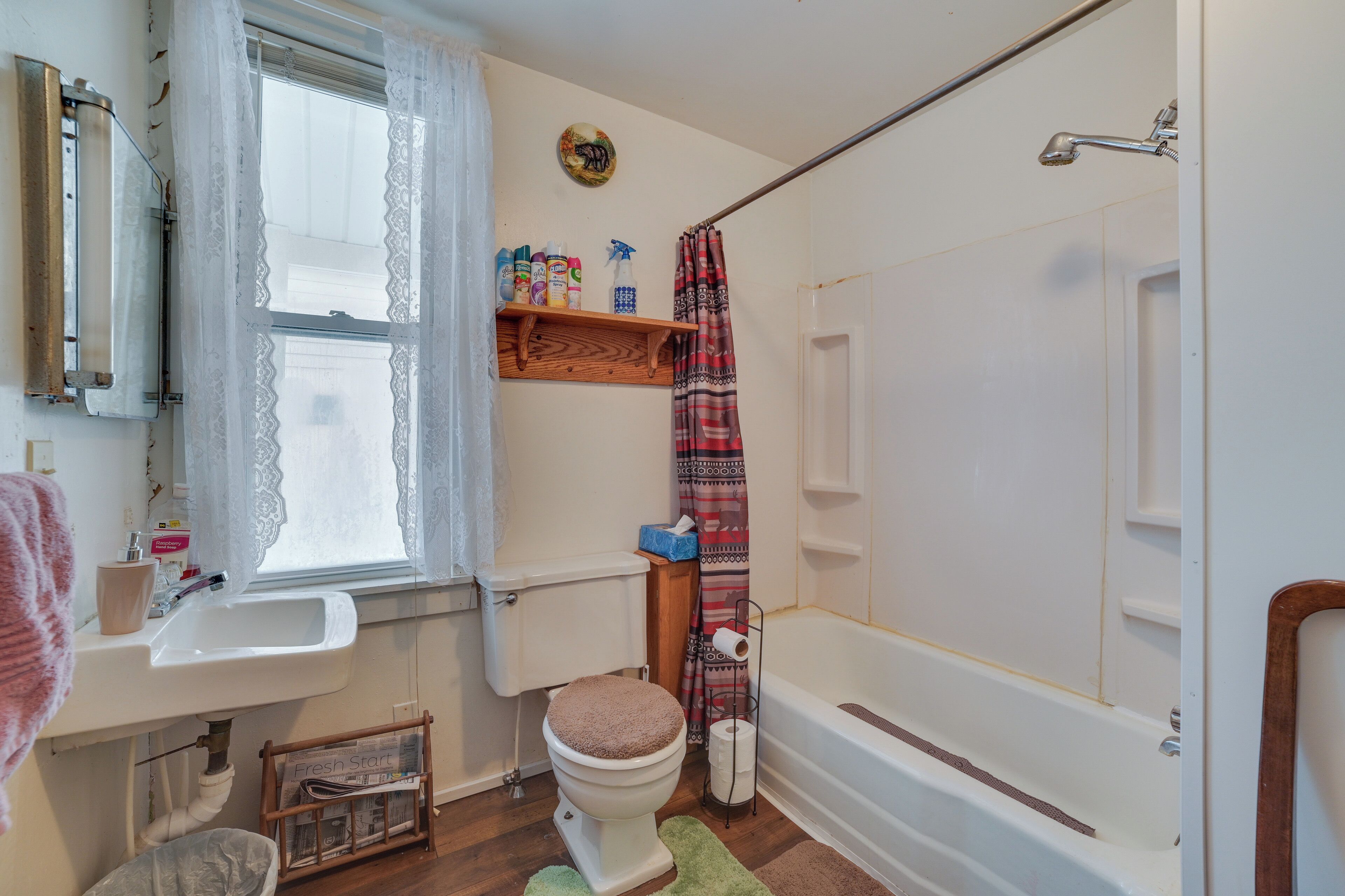 A bathroom with a toilet, sink, and bathtub. There is a window with lace curtains, a shower curtain, and a shelf with toiletries.