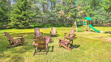 Relax in a semi-circle of Adirondack chairs around a central fire pit, surrounded by lush greenery and a family-friendly play structure.