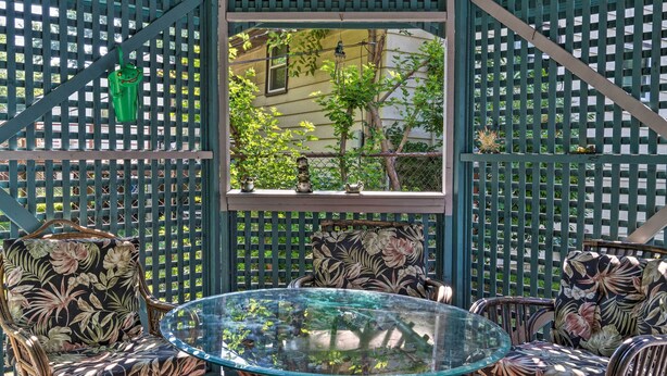 A glass-top table surrounded by wicker chairs with floral upholstery, set against a lattice backdrop with a window view of greenery. - Home w/ Fire Pit - 8 Mi to Dtwn Mile Square! (Indianapolis)