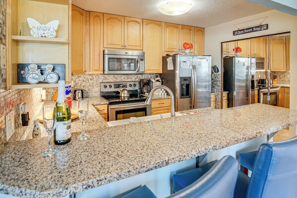A kitchen with granite countertops, stainless steel appliances, wooden cabinets, and a bar area with blue chairs. - < 1 Mi to Beach: Condo w/ Community Perks in Hhi! (Hilton Head Island)