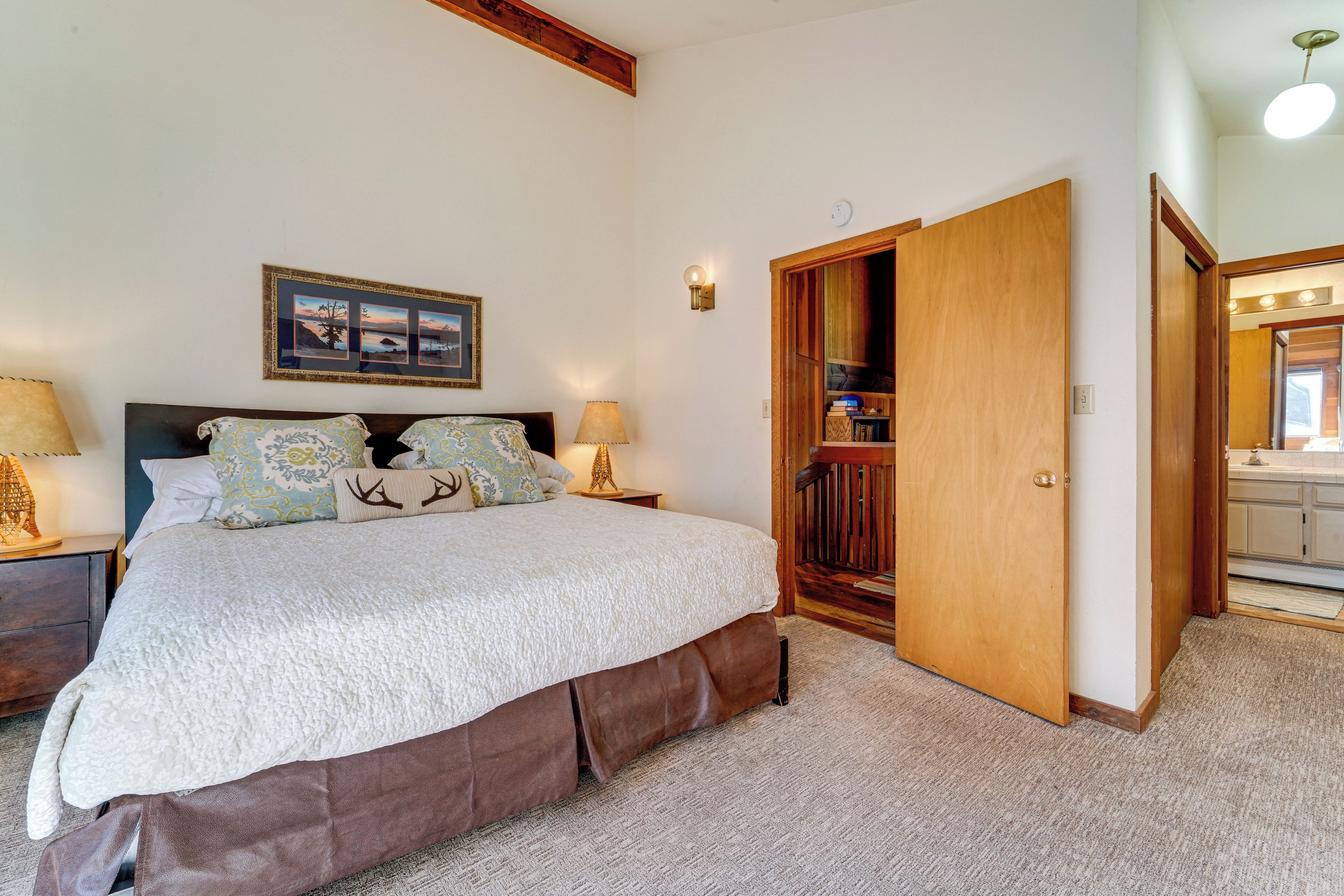 A bedroom with a large bed, a nightstand, a lamp, a wooden door, and a wall-mounted picture.