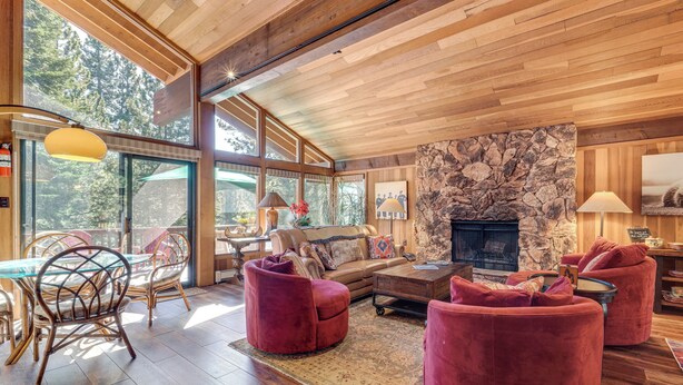 A spacious living room with a stone fireplace, wooden ceiling, and large windows offering a view of the forest. - Family-friendly Truckee Cabin Near Ski Shuttle! (Truckee)