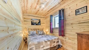 Une chambre dans une cabane en bois, avec un lit, une table de chevet, une fenêtre avec des rideaux et un tableau accroché au mur.