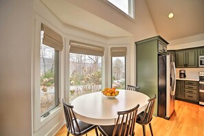 A dining area with a round table, chairs, and a fruit bowl. There are large windows with blinds, a kitchen with green cabinets, and a refrigerator. - Sapphire 5BR Luxe: 5 Private Baths, Mtn Views (Sapphire)