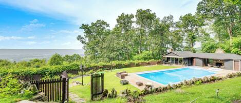 A backyard with a swimming pool, a patio with outdoor furniture, and a view of the surrounding landscape.