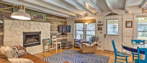A cozy living room with a stone fireplace, a wooden ceiling, and a dining area with blue chairs.