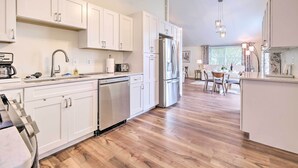 A modern kitchen with white cabinetry, a stainless steel refrigerator, and a dishwasher. There is a dining area with a table and chairs in the background. - Waterfront Cottage w/ Private Beach + Deck! (Colonial Beach)