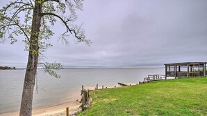 A lakeside view with a wooden deck, a grassy area, and a building near the water. - Waterfront Cottage w/ Private Beach + Deck! (Colonial Beach)