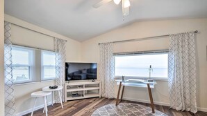 A living room with a TV, two white stools, a wooden floor, a patterned rug, and a view of the sea through a window. - Waterfront Cottage w/ Private Beach + Deck! (Colonial Beach)
