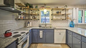 A modern kitchen with grey cabinets, a built-in stove, and a dishwasher. There are wooden shelves above the sink with various items and a window with a view of trees. - Renovated Eureka Springs Home - 1 Mi to Downtown! (Eureka Springs)