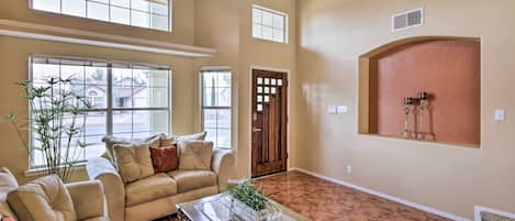 A spacious living room with a high ceiling, large windows, a ceiling fan, and a red-tiled floor.