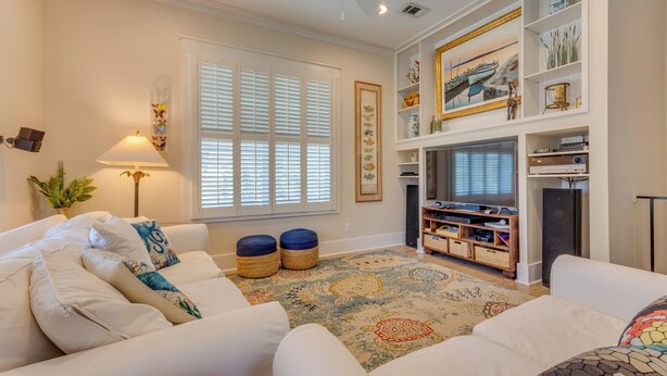 A living room with a white sofa, ottomans, a television, and a bookshelf. - Santa Rosa Beach Home w/ Pool, Hot Tub & Deck! (Santa Rosa Beach)