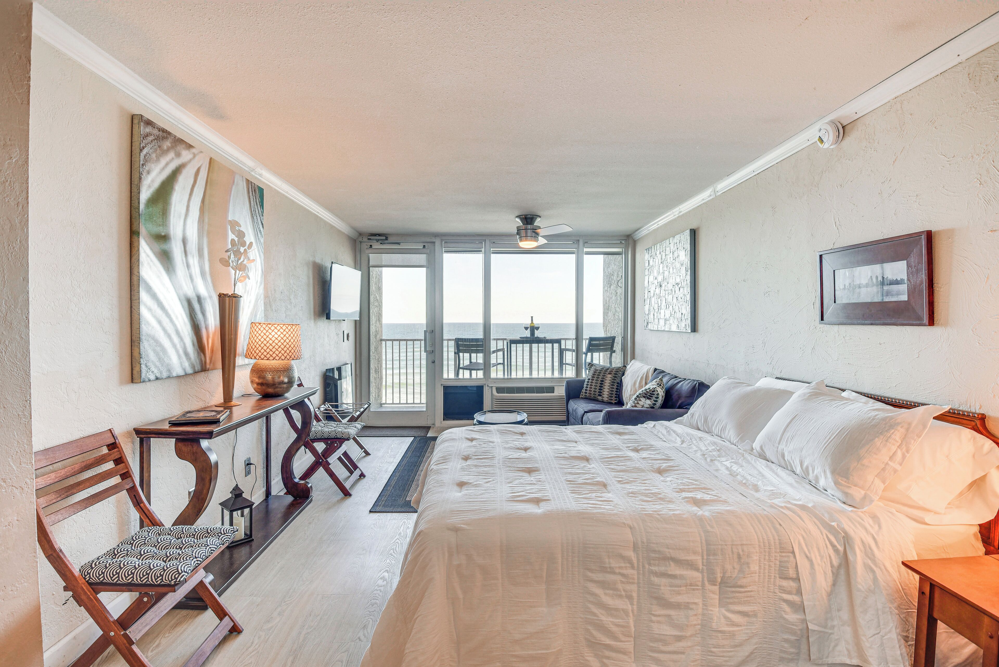 A hotel room with a large bed, a desk with a lamp, a chair, and a view of the sea through large windows.