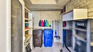 A compact utility room with a refrigerator, trash bins, and a broom rack against a brick wall. - Furnished Patio + Pool Access: Palm Springs Condo (Palm Springs)