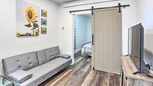Een moderne kamer met een grijze bank, een televisie op een houten standaard, een schuifdeur, en een muur met een zonnebloemschilderij en foto's.