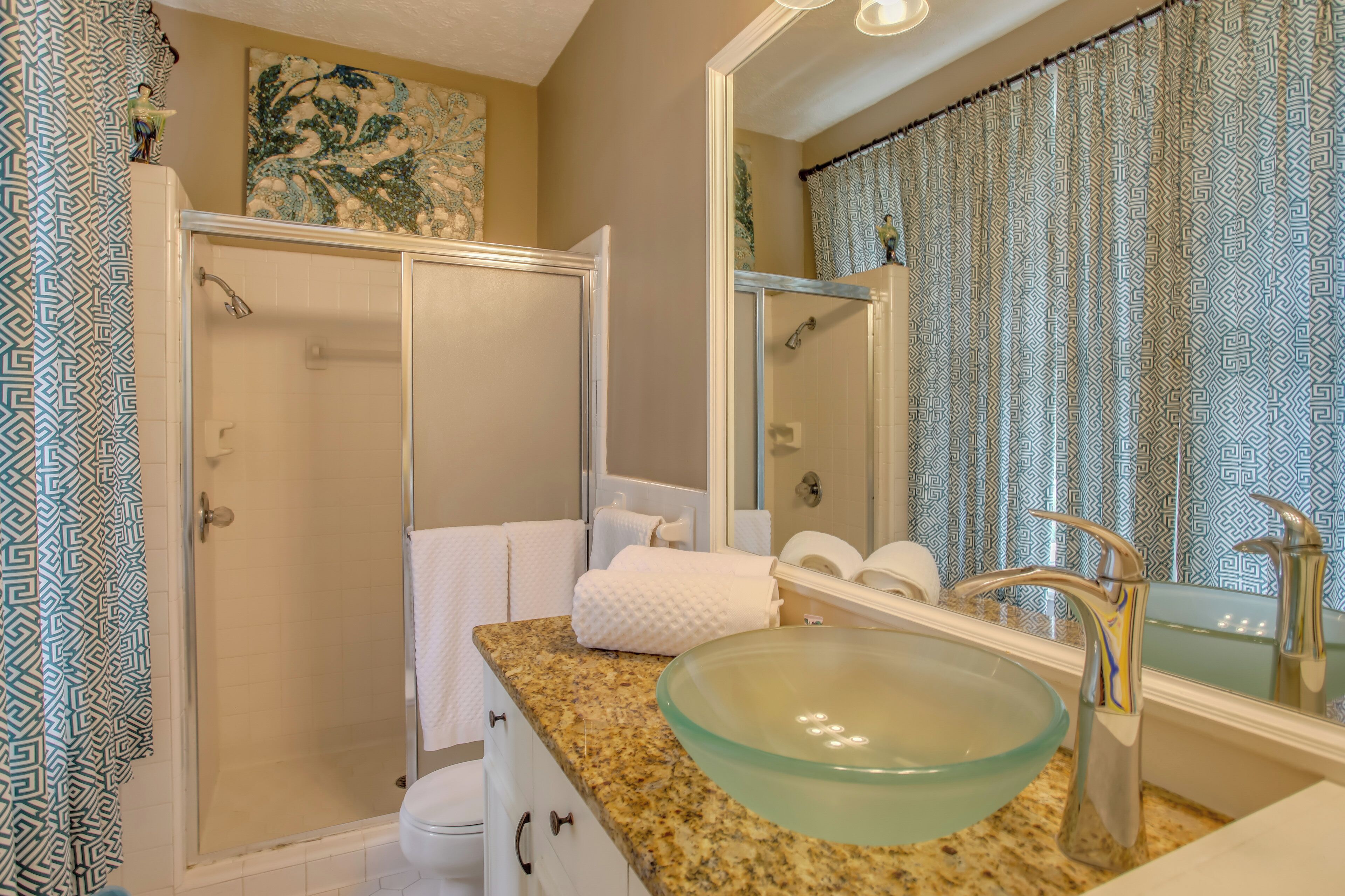 A bathroom with a glass bowl sink, a shower, and a mirror with a patterned curtain.