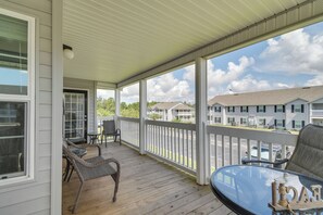 A covered outdoor area with a wooden deck, wicker furniture, and a glass table.