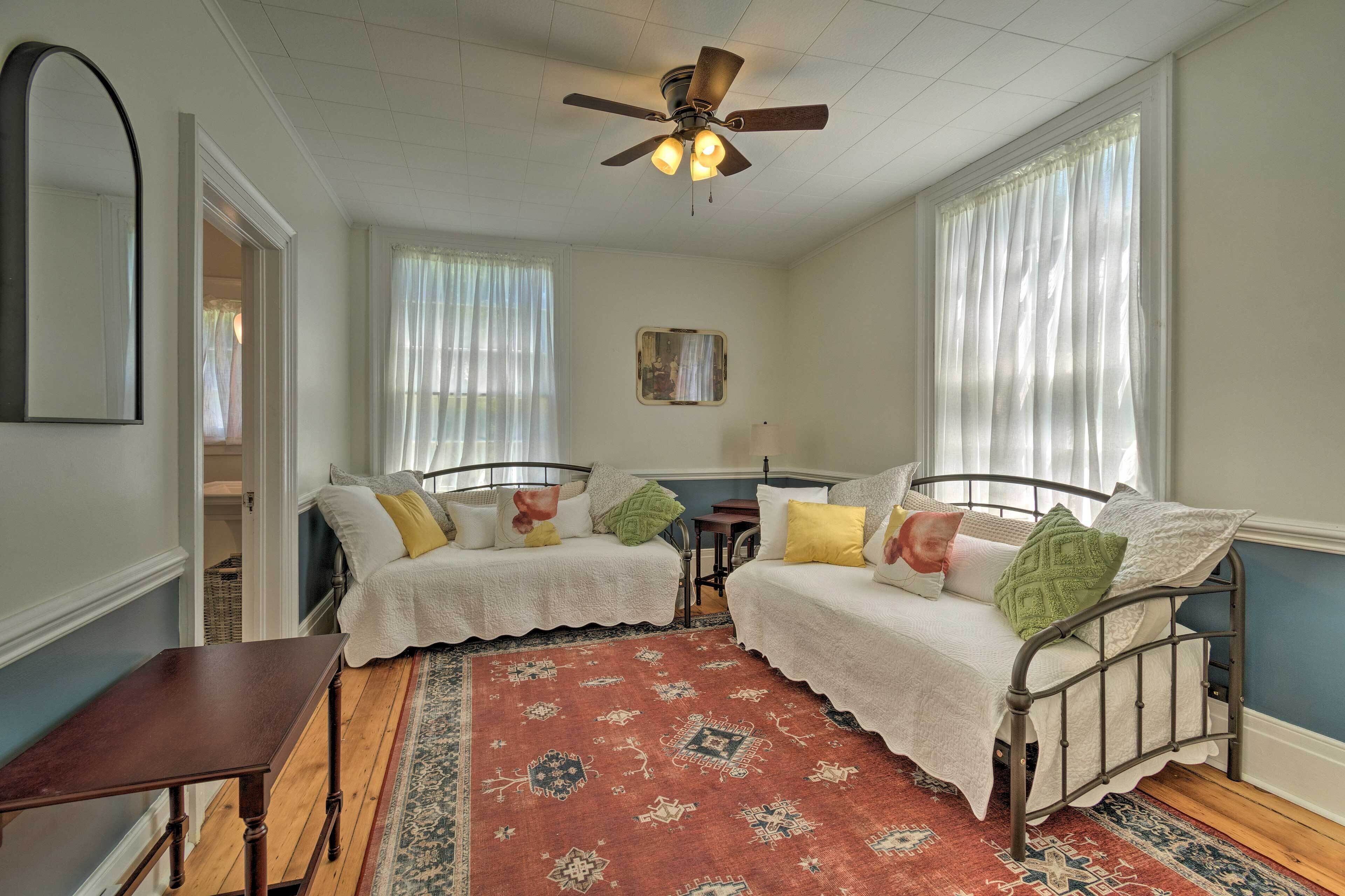A bedroom with two single beds, a ceiling fan, a large window with sheer curtains, a patterned rug, and a small table.