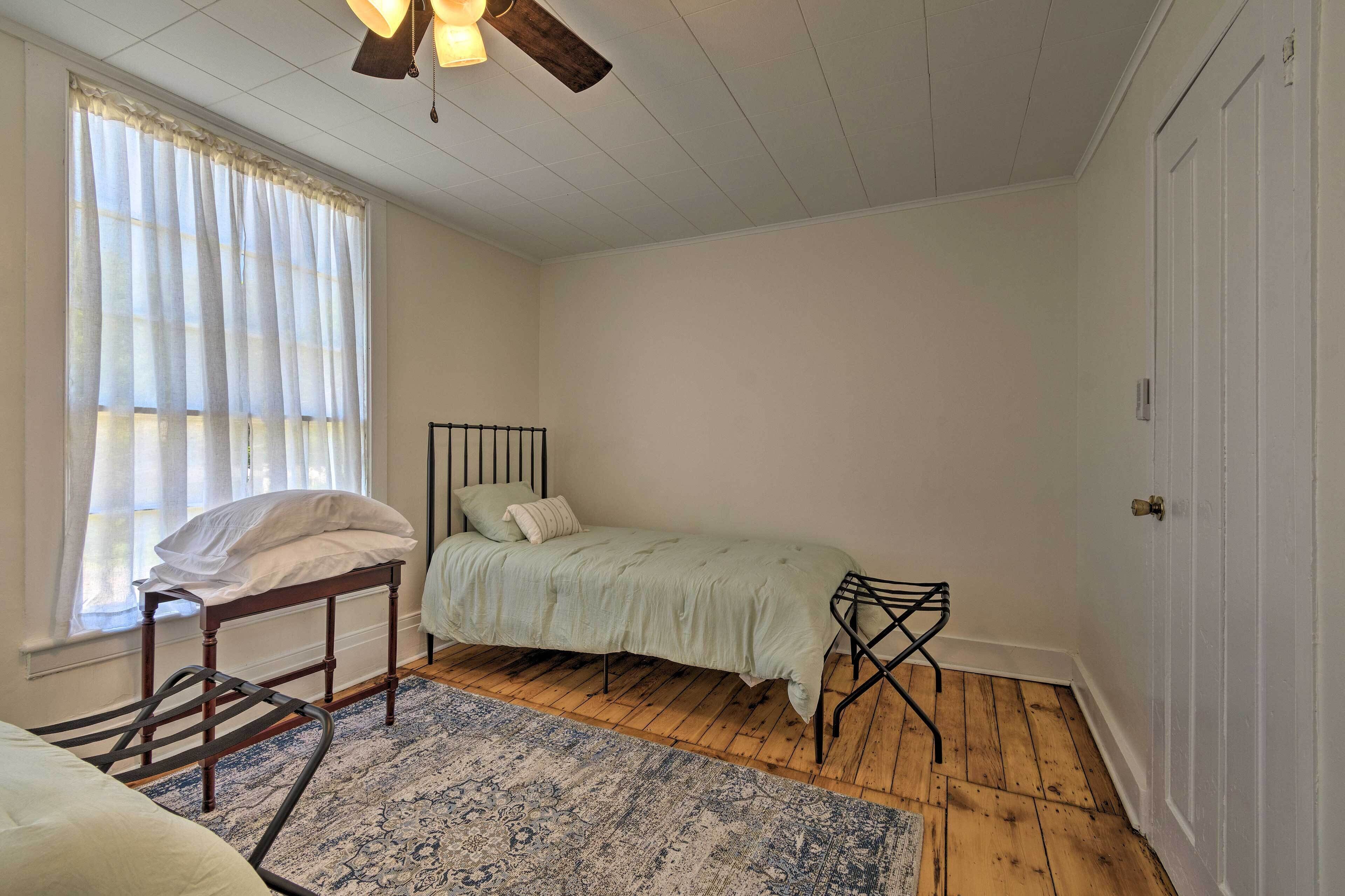 A bedroom with a single bed, a nightstand, a chair, a window with sheer curtains, and a ceiling fan.