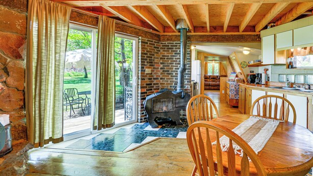 A rustic interior with a stone wall, wooden ceiling beams, a central wood-burning stove, a dining area with a table and chairs, and a kitchen with cabinets and a countertop. - 'snowflake Cabin' - Mins to Cody & Red Lodge! (Powell)