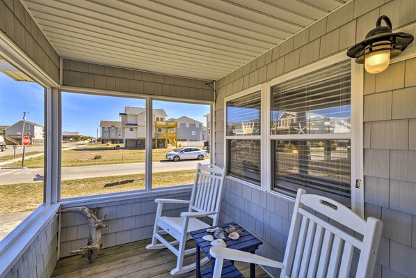 A covered porch with two rocking chairs and a small table, overlooking a street with houses and a car. - Nags Head Cottage: Screened Porch, Walk to Beach! (Nags Head)