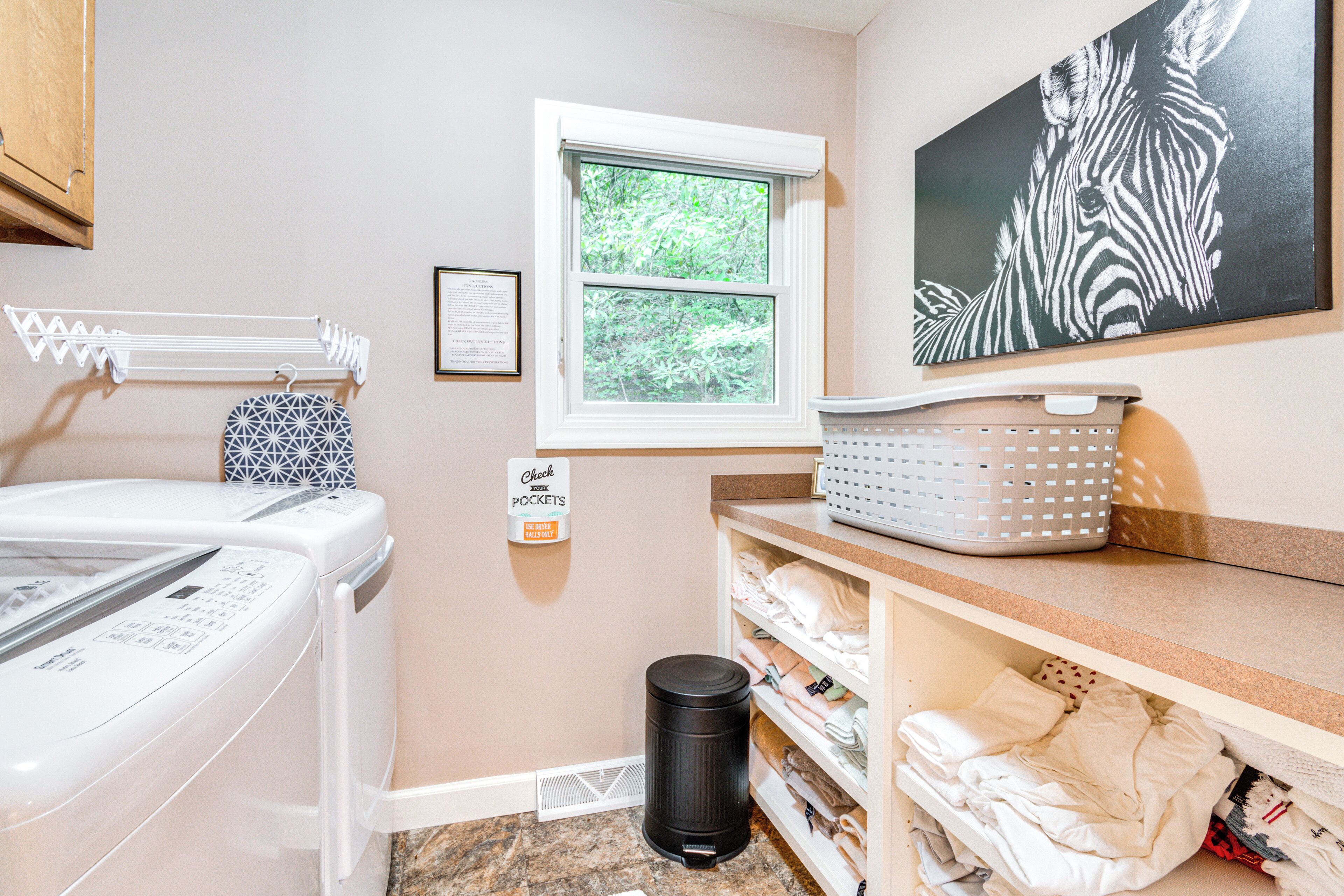 A laundry area with a washing machine, a drying rack, a trash bin, a window with a view of greenery, and a shelf with folded clothes.