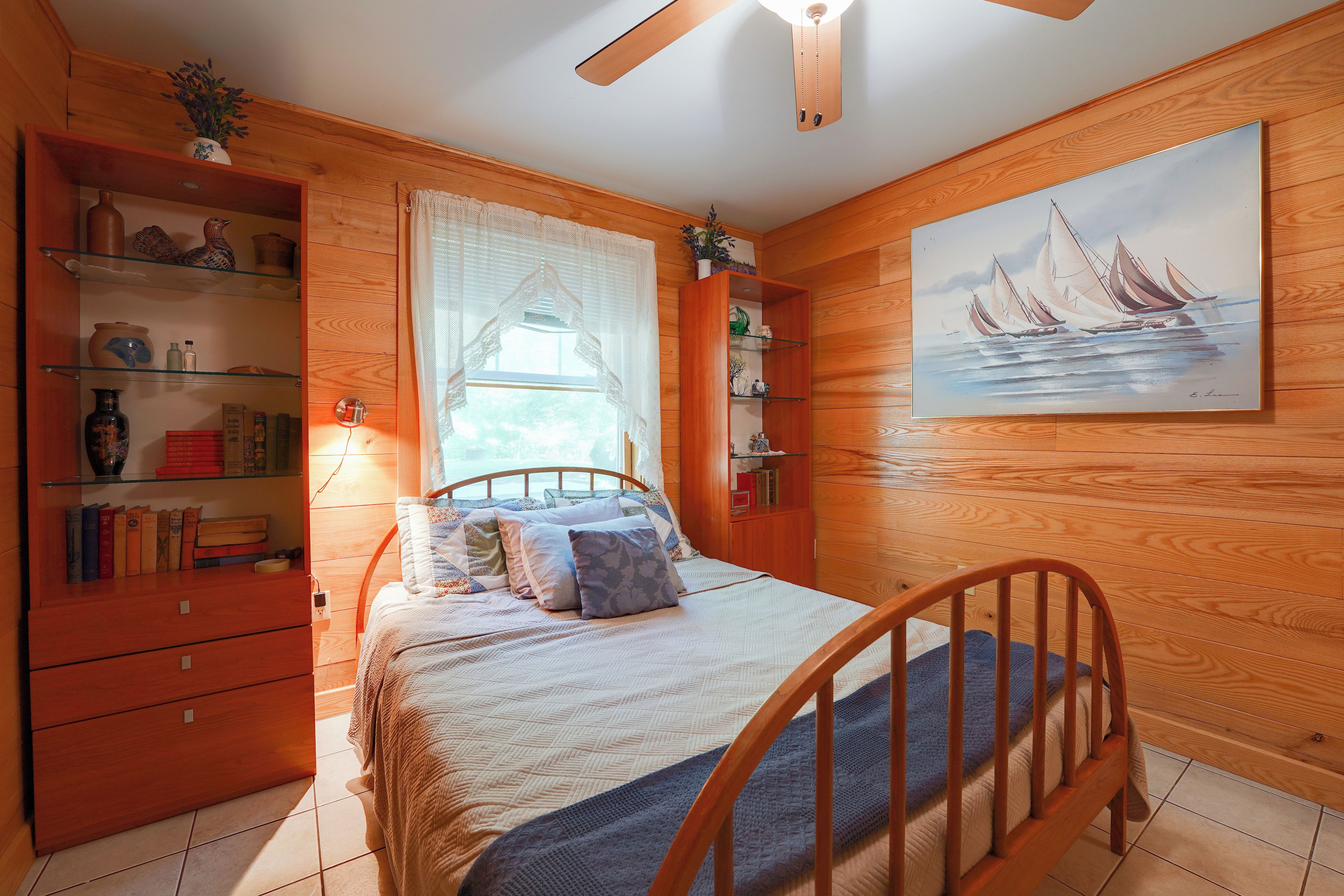 A bedroom with a wooden headboard, a bed with a quilt and pillows, a nightstand, a bookshelf, and a painting on the wall.