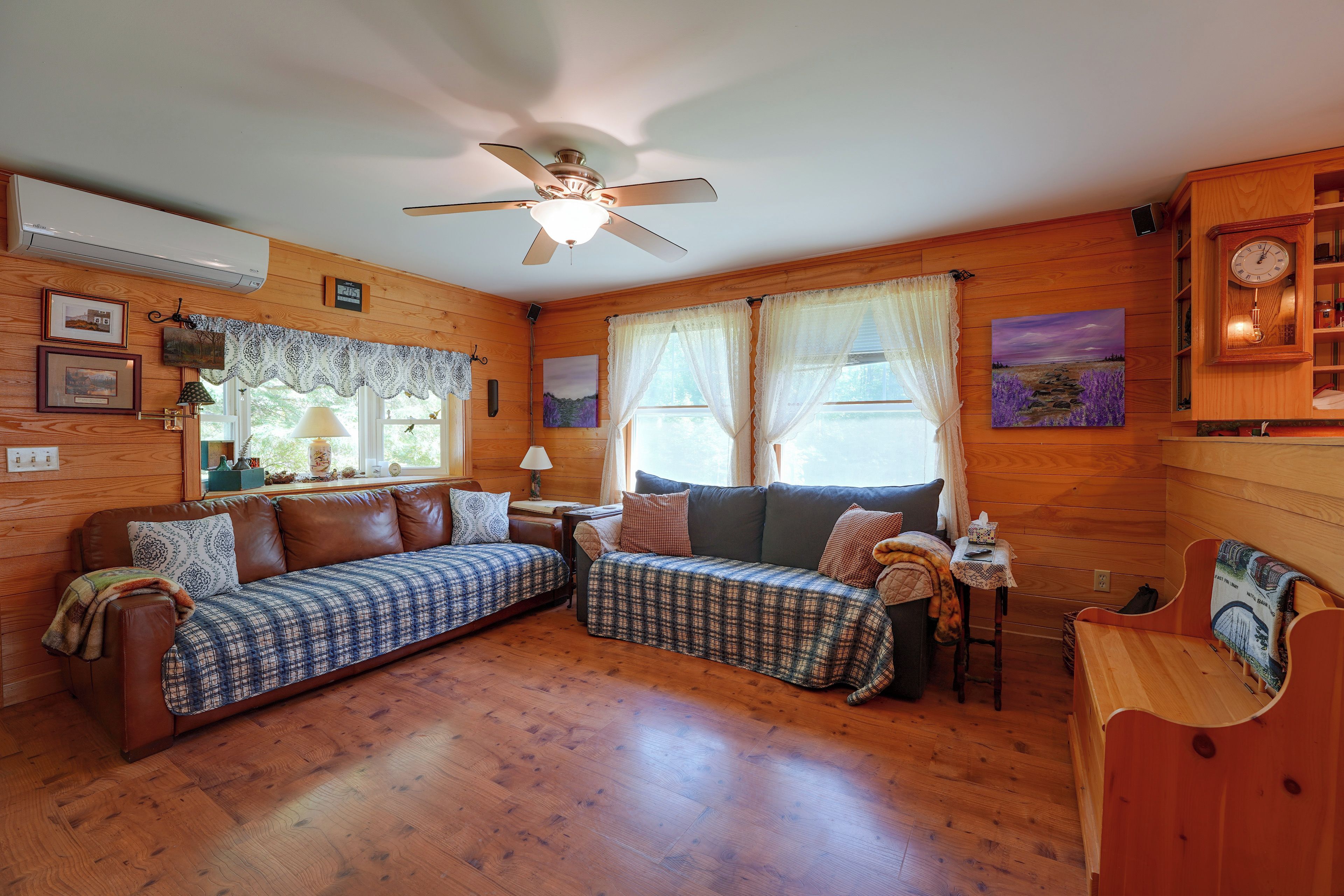 A cozy cabin interior with a wooden floor, a ceiling fan, a sofa, a small table, and a clock on the wall.